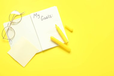 Listesi boş bir defter, sarı arka planda kalemler ve gözlükler. Yeni yıl hedefleri
