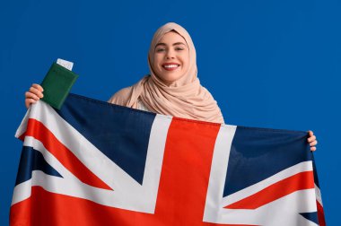 İngiliz bayrağı, pasaportu ve mavi arka planda bileti olan güzel, mutlu, genç bir Müslüman kadın.