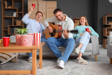 Arkadaşlarıyla evde gitar çalan genç bir adam.