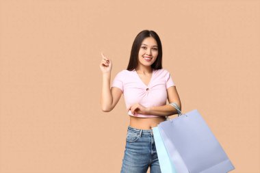 Kahverengi arka plandaki bir şeyi işaret eden alışveriş torbalarıyla şık giyinmiş şık Asyalı bir kadın.