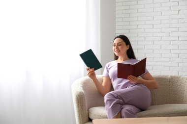 Kanepede oturan ve oturma odasında kitap tutan genç bir kadın.