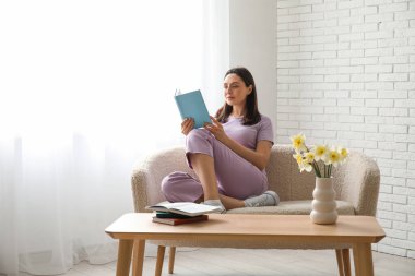 Genç bir kadın kanepede oturuyor ve oturma odasında kitap okuyor.