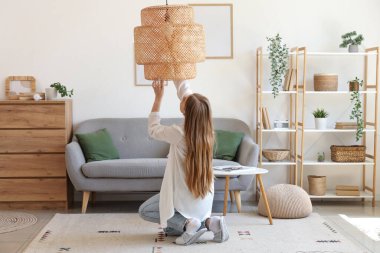 Ampulü vidalayan genç bir kadın evdeki, arkadaki lambaya takıyor.