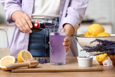 Mutfaktaki masada limonata hazırlamak için şişeden cama lavanta suyu döken genç bir kadın.