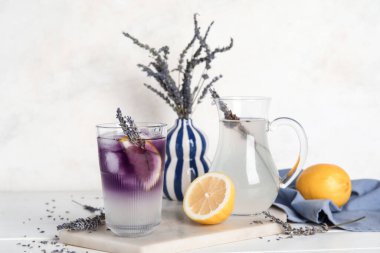 Bir bardak soğuk lavantalı limonata, limonlar ve çiçekler açık ahşap arka planda.