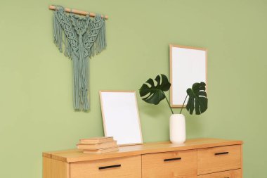 Yeşil duvarın yanındaki ahşap dolapta canavar yaprakları, kitaplar ve çerçeve var.