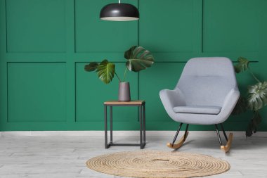 Yeşil duvarın yanındaki kahve masasında canavar yaprakları olan gri koltuk ve vazo.