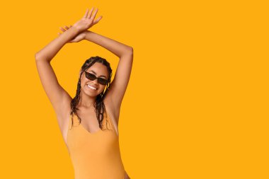 Sarı arka planda şık mayolu ve güneş gözlüklü güzel Afro-Amerikan kadın.