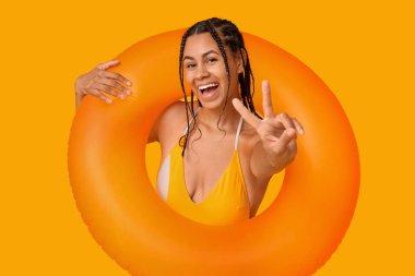 Sarı arka planda zafer jesti gösteren şişirilebilir yüzüklü şık mayolu güzel Afro-Amerikalı kadın.