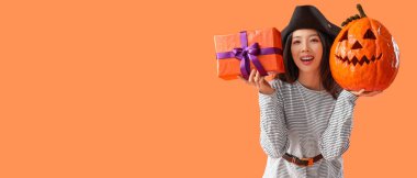 Korsan gibi giyinmiş genç Asyalı kadın elinde Cadılar Bayramı balkabağı ve turuncu arka planda mesaj için yeri olan bir hediye tutuyor.