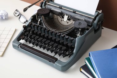 Kahverengi duvarın yanındaki beyaz masada defter, gözlük ve kitaplarla klasik daktilo.