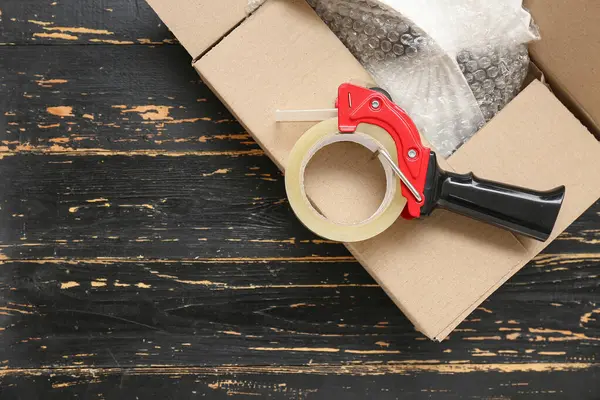 stock image Packing tape dispenser and cardboard box on black wooden background