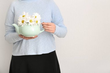 Beyaz duvarın yanında çaydanlık dolusu papatya çayı ve çiçeklerle güzel genç bir kadın.