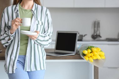 Masanın yanında modern laptop ve sarı lale çiçekleriyle kahve içen bir kadın.