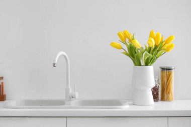 Mutfak lavabosunun yanındaki tezgahta sarı lale çiçekleri olan vazo.