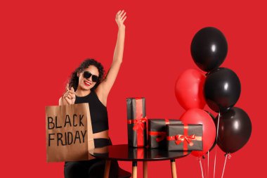 Kırmızı arka planda oturan hediye kutuları, balonlar ve alışveriş çantasıyla güzel bir Afro-Amerikalı kadın. Kara Cuma indirimi