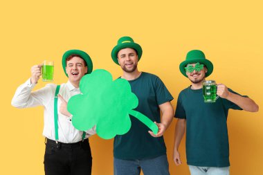 Sarı arka planda bira ve yonca taşıyan cüce şapkalı mutlu genç adamlar. Aziz Patrick Günü kutlaması