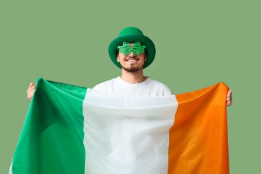 Yeşil arka planda İrlanda bayrağı ve parti gözlükleriyle cüce şapkalı mutlu genç adam. Aziz Patrick Günü kutlaması