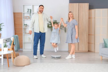 Evde annesiyle babası robot süpürgenin üzerinden atlayan küçük bir kız.