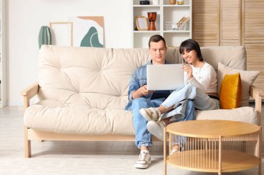 Genç çift evdeki kanepede dizüstü bilgisayar kullanıyor.