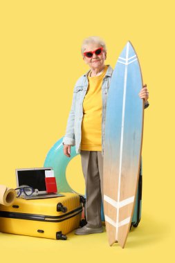 Sarı arka planda sörf tahtası ve seyahat aksesuarları olan son sınıf bayan turist.