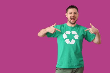 Mutlu genç adam tişörtündeki geri dönüşüm logosunu pembe arka planda gösteriyor. Ekoloji kavramı