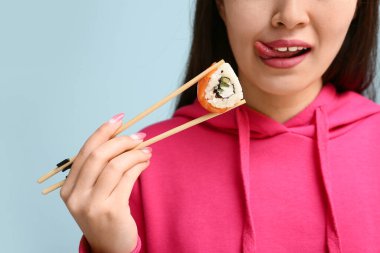 Lezzetli suşi rulosu olan güzel genç bir kadın mavi arka planda dilini gösteriyor, yakın plan.