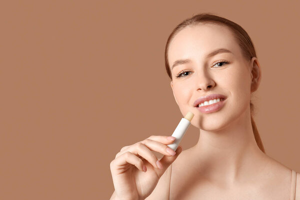 Beautiful young woman applying lip balm on brown background