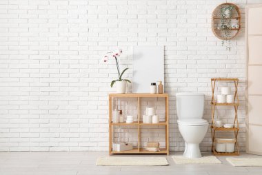 Beyaz tuğla duvarın yanında banyo aksesuarları olan klozet ve raf ünitesi.