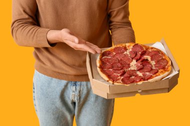 Sarı arka planda lezzetli pepperonili pizza olan karton kutu tutan genç adam, yakın plan.