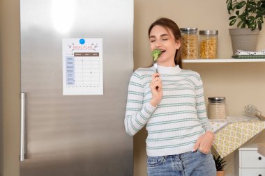 Mutfakta buzdolabının yanında brokoli yiyen genç bir kadın. Diyet kavramı