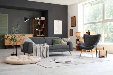 Modern oturma odasının içinde siyah kanepe, dekoratif zeytin ağacı ve yerde dizüstü bilgisayar.