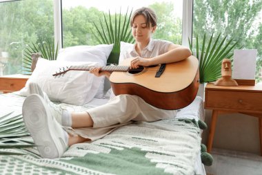 Genç bir kadın yatak odasında gitar çalıyor.