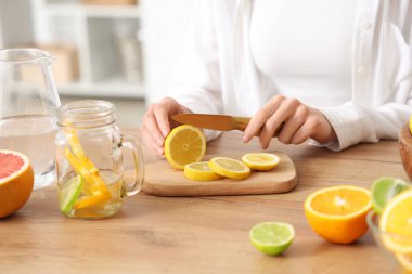 Mutfakta eritilmiş su hazırlamak için farklı narenciye meyveleri kesen genç bir kadın.