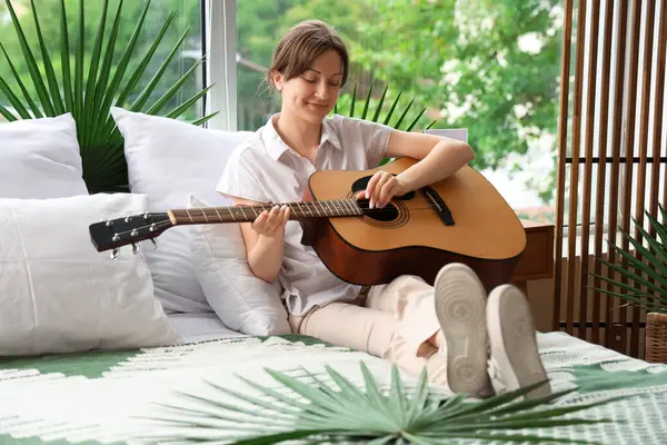 Genç bir kadın yatak odasında gitar çalıyor.