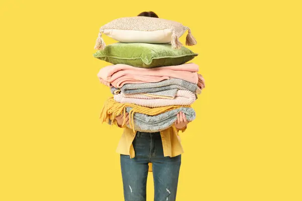 stock image Beautiful young woman with stack of different blankets and pillows on yellow background