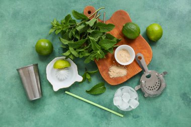 Ahşap kesme tahtası, çalkalayıcı, süzgeç ve yeşil arka planda mojito malzemeleri.