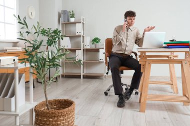 Erkek muhasebeci cep telefonuyla konuşuyor ve işyerinde dizüstü bilgisayar kullanıyor.