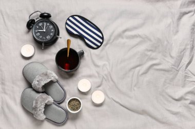 Terlikler, çalar saat, uyku maskesi, mumlar ve gri kumaş arka planda bir fincan çay.