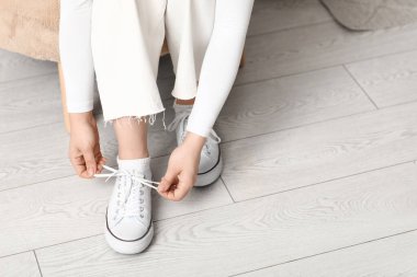 Spor ayakkabılı ve beyaz pantolonlu genç bir kadın evde ayakkabı bağı bağlıyor.