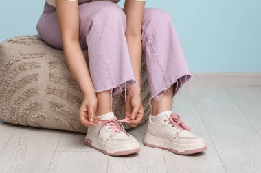 Şık ayakkabılı ve kot pantolonlu bir kadın mavi arka plana pembe ayakkabı bağlıyor.