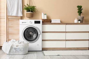 Evdeki bej duvarın yanında şifonyer ve çamaşır sepetli modern çamaşır makinesi.