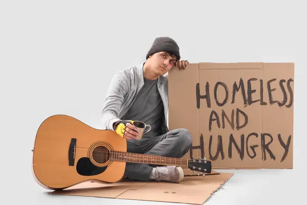 stock image Young homeless man with cup and guitar begging for money on light background