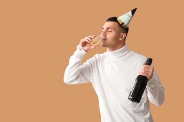 Parti şapkalı, elinde şişe ve bir bardak şampanyayla genç adam kahverengi arka planda doğum gününü kutluyor.