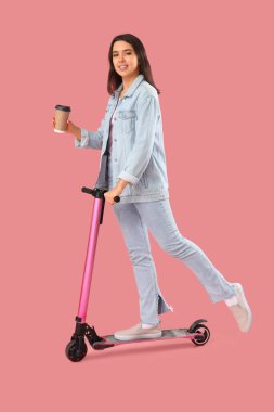 Kahve fincanı olan genç bir kadın pembe arka planda elektrikli scooter kullanıyor.