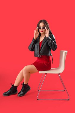 Şık giyinmiş, güneş gözlüklü güzel bir Asyalı kadın kırmızı arka planda, sandalyede oturuyor.