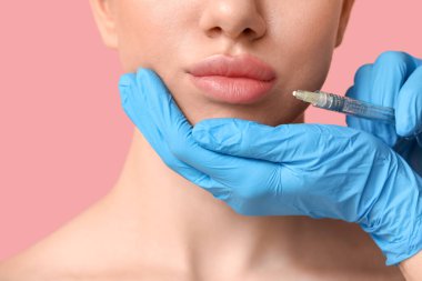 Genç bir kadın pembe arka planda dudaklarından enjeksiyon yaptırıyor, yakın plan.
