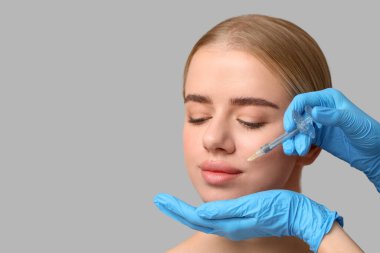 Gri arka planda dudaklarından doldurucu enjeksiyon alan genç kadın.