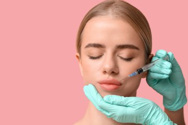 Genç bir kadın pembe arka planda dudaklarından enjeksiyon yaptırıyor.