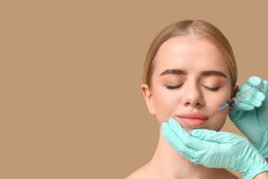 Genç bir kadın kahverengi arka planda dudaklarından enjeksiyon yaptırıyor.